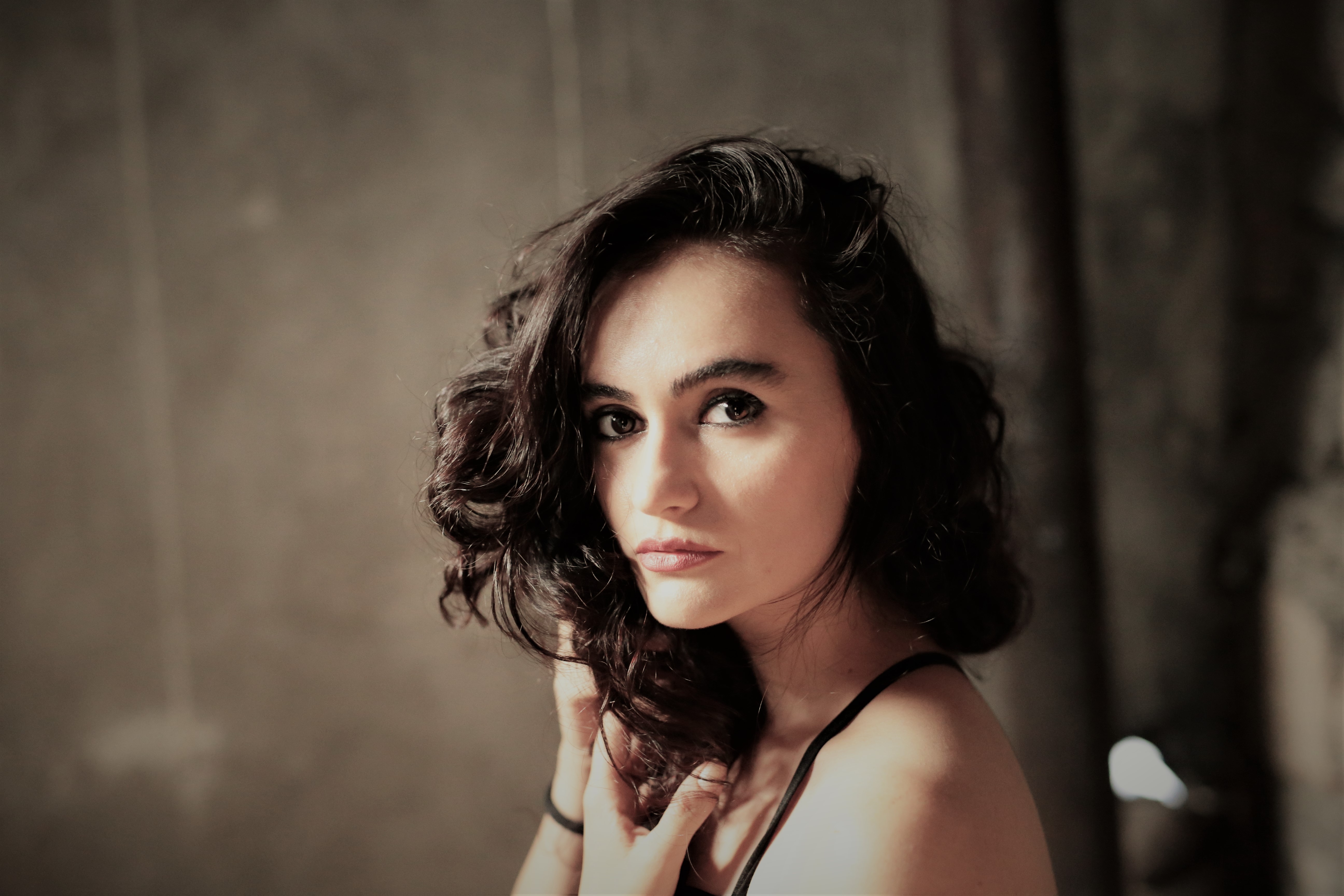 A woman with long, wavy hair looks directly at the camera with a thoughtful expression, set against a blurred concrete background.